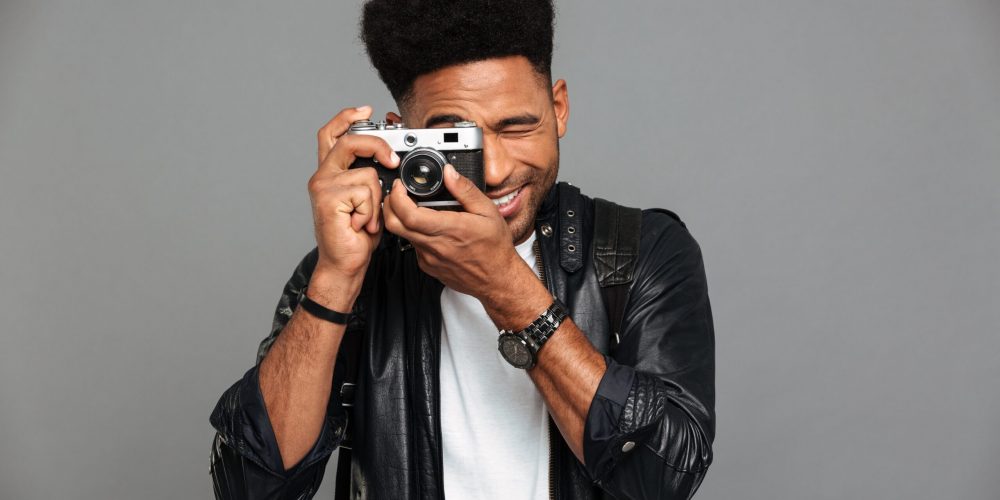 young-cheerful-african-man-looking-through-retro-cameras-objective-while-taking-photo-min